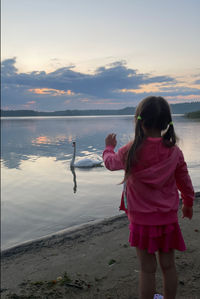 A girl standing at sunset on a lake shore saying goodbye to a swan. 