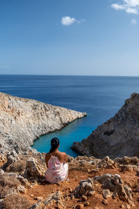 Rear view of woman sitting on rock by sea against sky