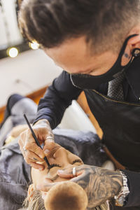 High angle of tattooed man in mask drawing eyeliner arrows on eyelids of woman during work in makeup studio