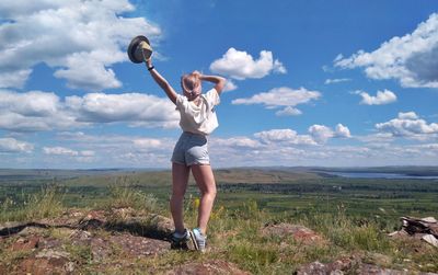 Rear view full length of woman holding hat on landscape against sky