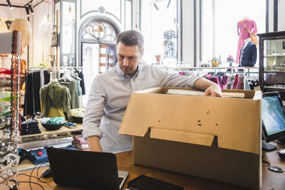 Mature entrepreneur using laptop while unpacking cardboard box in boutique