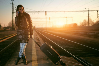 Young woman with luggage standing at railroad station platform against sky during sunset