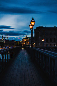 Illuminated street light on footbridge in city against sky at dusk