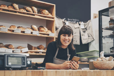 Smiling store owner with mobile phone leaning on checkout counter in shop