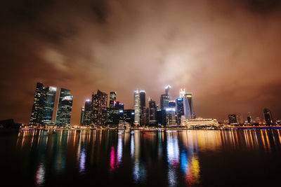 Illuminated modern buildings by bay against sky at night