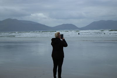 Rear view of woman photographing sea while standing at beach against sky