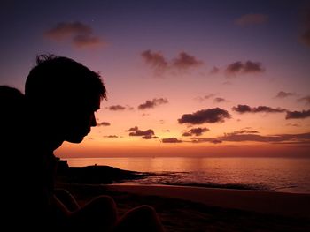 Man sitting at beach against sky during sunset