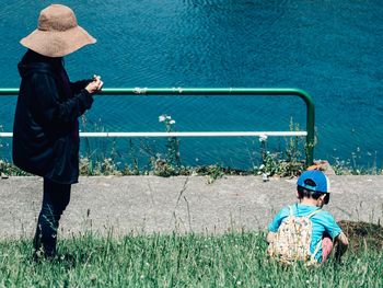 Side view of mother standing by son crouching on grassy field by lake