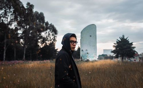 Portrait of young man wearing hoodie standing on field against sky