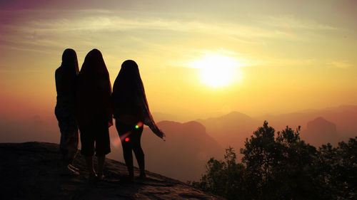 Friends standing on rock by fog covered mountains against orange sky