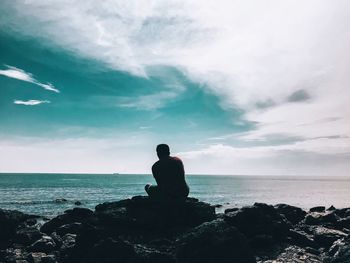 Man sitting on rock looking at sea against sky