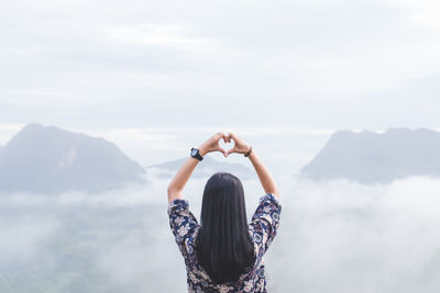 Rear view of woman making heart shape while looking at mountains