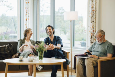 Cheerful senior woman talking with male nurse sitting on sofa against window at nursing home