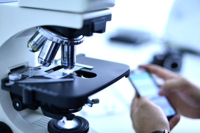 Cropped hands of male scientist using mobile phone by microscope at laboratory