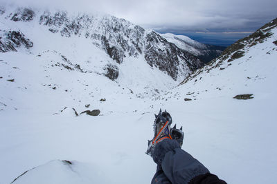 Low section of man sitting on snowcapped mountain against cloudy sky