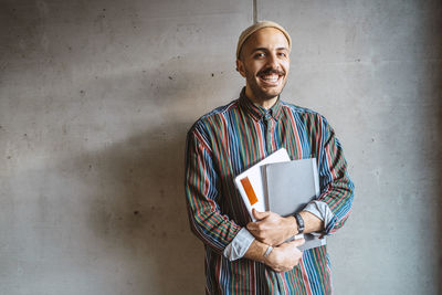 Portrait of cheerful male adult student holding book standing in front of gray wall at college