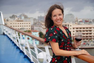 Portrait of smiling young woman standing by railing in city against sky