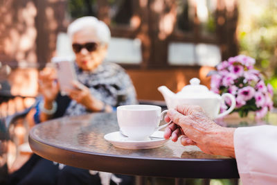 Senior man hand holding a cup of tea with his wife in background.