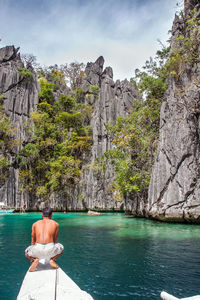 Rear view of shirtless man crouching on boat by cliff against sky