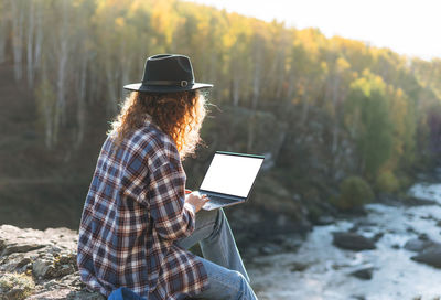Young woman with curly hair in felt hat and plaid shirt using laptop on view of mountains and river