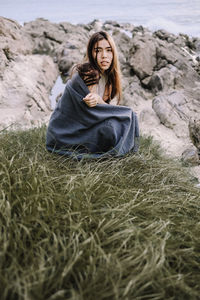 Portrait of woman sitting on field against rocks at beach