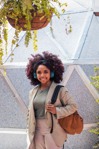 Optimistic young african american female manager in suit with backpack looking at camera with smile and listening to music while standing against geometric wall with potted plants on sunny day