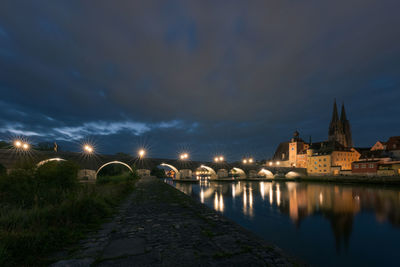 Bridge over river by illuminated buildings against sky at night