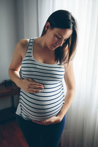 Pregnant young woman standing against window at home