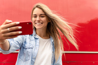 Portrait of smiling young woman taking selfie standing against red background