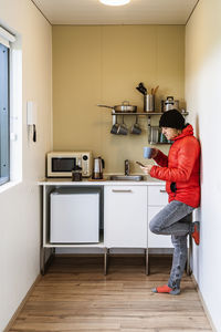 Side view of young male traveler in warm jacket standing in kitchen leaning on wall while drinking cup of hot coffee and messaging on mobile phone