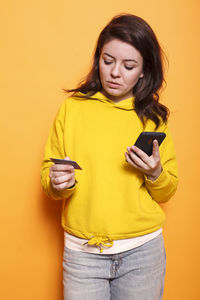 Young woman using mobile phone while standing against yellow background