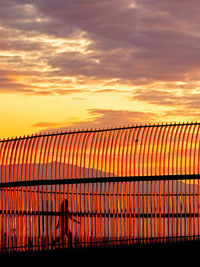 Silhouette person walking behind fence against orange sky