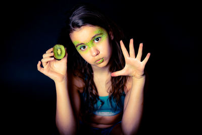 Portrait of beautiful young woman with face paint holding kiwi against black background