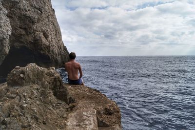 Rear view of shirtless man sitting on rock against sea at beach