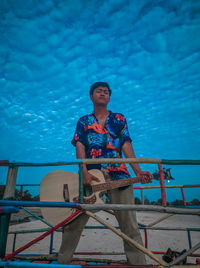 Young man sitting on railing against blue sky