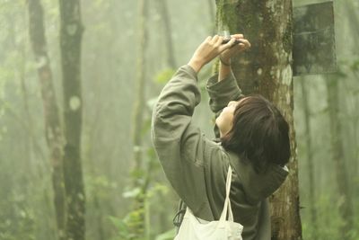 Woman photographing through camera while standing by tree at forest