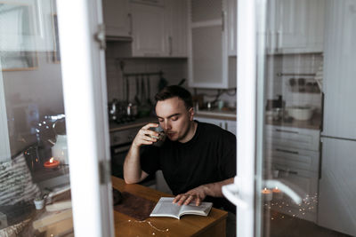 Young man drinking tea and reading a book in his modern light kitchen