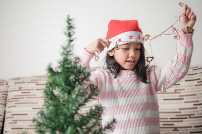 Cute girl with arms raised holding christmas decoration while standing against wall