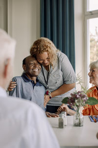 Smiling young female caregiver embracing happy senior men sitting with friends at dining table in nursing home