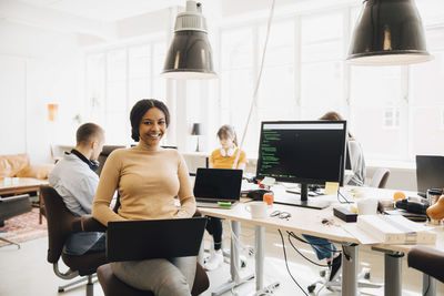 Portrait of smiling female programmer using laptop while coworkers working in background