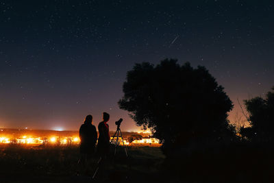 Silhouette female friends standing with telescope on field against star field