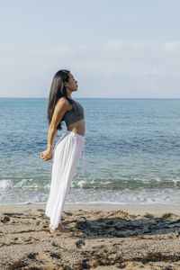 Side view of ethnic female leaning back while standing in ashta chandrasana pose during yoga practice on sandy coast against ocean