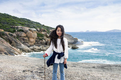 Portrait of young woman standing on rock at beach against sky