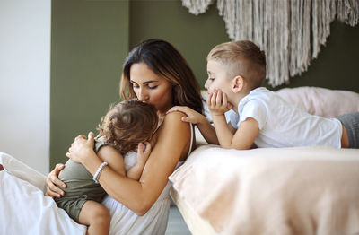 Mother embracing daughter while sitting by bed at home