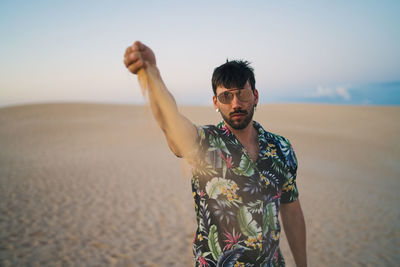 Young man wearing sunglasses standing on sand at beach against sky