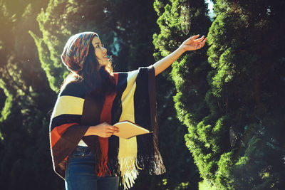 Side view of woman with book and pen touching tree on land