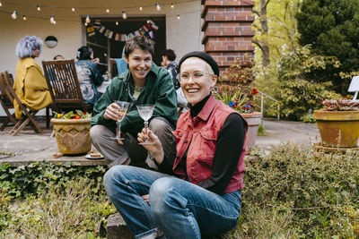 Smiling gay women sitting together holding wineglasses near plants