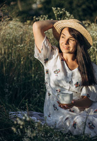 A girl in a straw hat sits in nature with a cup in her hands and smiles