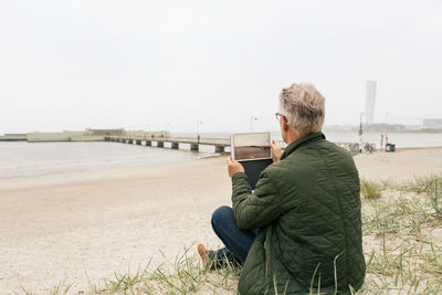 Rear view of senior male commuter photographing sea while sitting on sand at beach against clear sky
