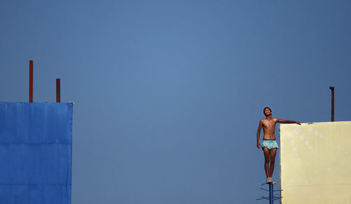 Shirtless young man standing on pole against clear blue sky